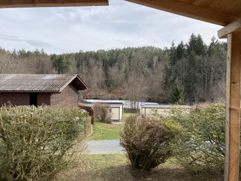 View from Stuga 2 at Wörthersee, Austria, showing wooden lodges, shrubs, and a forested hillside nearby.