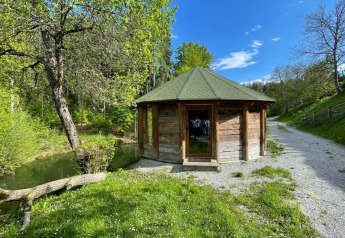 Lille træhus ved en grussti omgivet af frodig natur og grønne træer under en klar blå himmel.