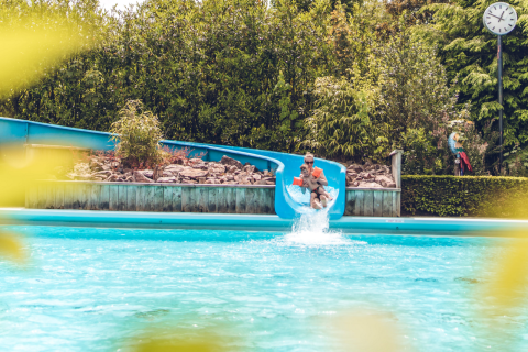 Padre e hijo bajan por un tobogán acuático hacia la piscina en un alojamiento glamping rodeado de vegetación.