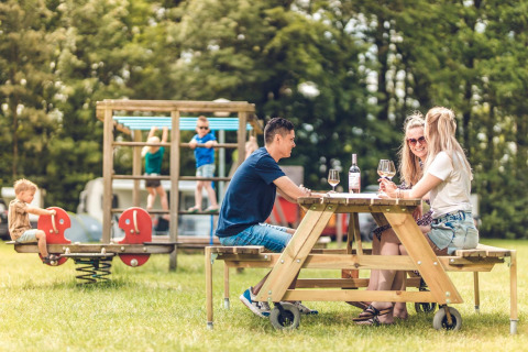 Adultos disfrutan vino en una mesa de picnic mientras niños juegan en un parque en un glamping.
