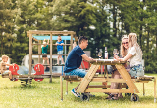 Adultos disfrutan vino en una mesa de picnic mientras niños juegan en un parque en un glamping.