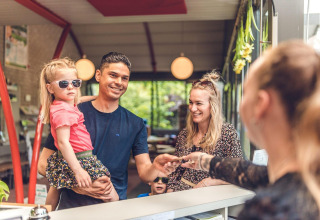 Familia registrándose en alojamiento glamping. Padre sostiene hija con gafas de sol ante recepcionista.