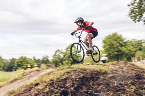 Una persona salta con bicicleta sobre un montículo de tierra en un alojamiento glamping rodeado de vegetación.