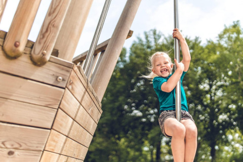 Niña sonriente deslizándose por un tubo de un parque de madera en un glamping con árboles verdes de fondo.