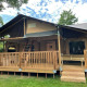 Safari telt kaldet Ranger Lodge med træterrasse, netloft og bænk, omgivet af grønne træer og græs.