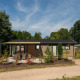 Veluwe Lodge set udefra, omgivet af træer og buske under en blå himmel med spredte skyer.