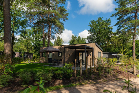 Moderna cabaña Loevestein en bosque, con terraza, mesa exterior, sombrilla y altos árboles verdes.