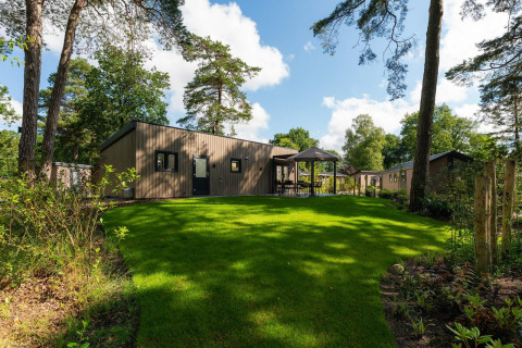 Moderne Holzlodge mit Rasen und überdachter Terrasse in grünem, waldreichen Naturgebiet bei Sonnenschein.