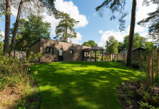 Moderne lodge met houten buitenkant, groen gazon en terras in boszone, onder blauwe lucht met zon.