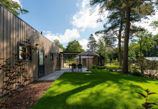 Lodge moderno con terraza, césped verde y altos árboles alrededor, bajo un cielo despejado y soleado.