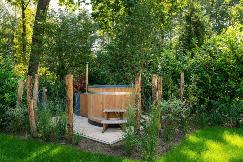 Bañera de hidromasaje de madera al aire libre rodeada de vegetación y postes rústicos en una cabaña.