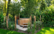 Buiten houten hottub omringd door groene planten en palen in de tuin bij een lodge in de zomer.