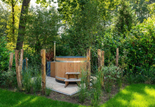 Bañera de hidromasaje de madera al aire libre rodeada de vegetación y postes rústicos en una cabaña.