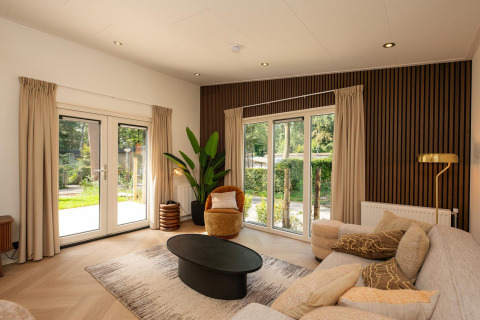 Sala de lodge moderna con grandes ventanales, sofá beige, planta verde y abundante luz natural.