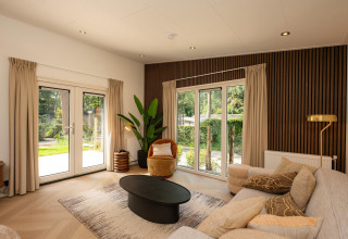 Sala de lodge moderna con grandes ventanales, sofá beige, planta verde y abundante luz natural.
