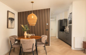 Moderne lodge-spisestue med rundt bord, fire polstrede stole, træpanelvæg og køkken i baggrunden.
