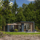 Moderne lodge med store vinduer, terrasse og havemøbler, omgivet af grønne træer og natur under blå himmel.
