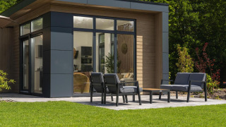 Moderne lodge met grote ramen, zithoek buiten met tuinmeubilair en groen gazon op de voorgrond.