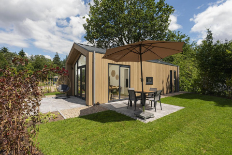 Moderna Tiny Lodge con patio, sombrilla y silla colgante en un jardín verde, cielo parcialmente nublado.