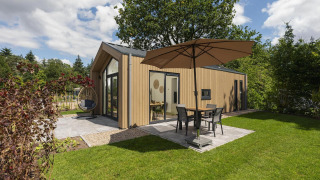 Modern Tiny Lodge with patio, umbrella, and hanging chair in a green garden, partly cloudy sky above.