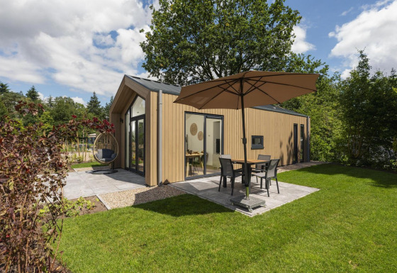 Moderna Tiny Lodge con patio, sombrilla y silla colgante en un jardín verde, cielo parcialmente nublado.