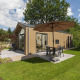 Moderne Tiny Lodge med terrasse, parasol og hængestol i en grøn have, himlen er delvist skyet.
