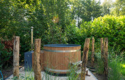 Buiten hottub van hout omgeven door natuur en palen, gefotografeerd bij een groene lodgeomgeving.