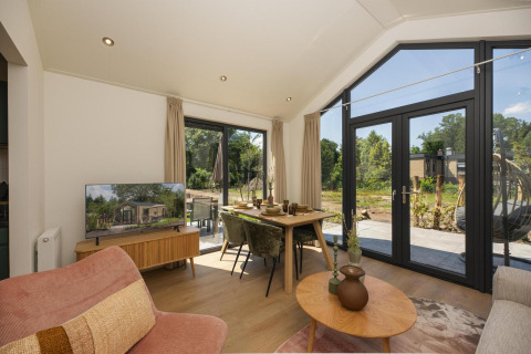 Bright living area with large windows, dining table, and TV in the Tiny Lodge with Hot tub at Veluwepark de Bosgraaf, Netherlands.