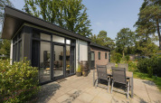 Udendørs terrasse ved Heather Lodge på Veluwepark de Bosgraaf i Holland, omgivet af grønne træer.