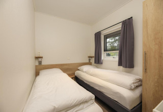 Kamer met twee eenpersoonsbedden, wit beddengoed, raam met grijze gordijnen in Heather Lodge, Veluwepark de Bosgraaf.