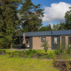 Velthorst Comfort lodge set i et grønt skovområde med moderne facade og udendørs siddeområde under en paraply.