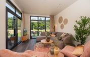 Moderne stue i en lodge ved Cannenburch Deluxe, Veluwepark de Bosgraaf, Holland, med store vinduer og stilfuld indretning.