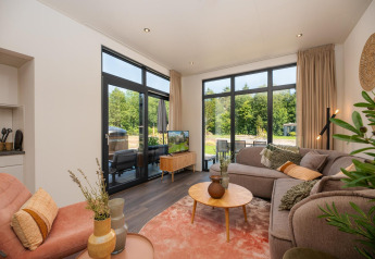 Stue med store vinduer, komfortabel sofa og udsigt til have i Cannenburch Deluxe-lodge, Veluwepark de Bosgraaf.