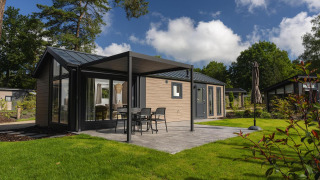 Lodge moderna con patio, mobili da esterno e ombrellone, circondata da prato verde e alberi alti.