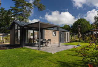 Lodge moderna con terraza, muebles de exterior y sombrilla, rodeada de césped verde y árboles altos.