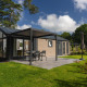 Moderne lodge med terrasse, havemøbler og parasol, omgivet af grønt græs og træer under blå himmel.