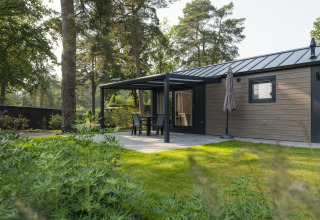 Cabaña moderna con terraza techada, muebles de jardín y sombrilla, rodeada de árboles y vegetación.