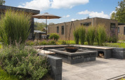 Outdoor lounge fire pit area at a modern wooden lodge, surrounded by tall grass, plants, and a parasol.