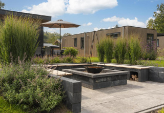 Zona de fogata en un lodge moderno de madera con bancos, plantas altas y una sombrilla en el jardín.
