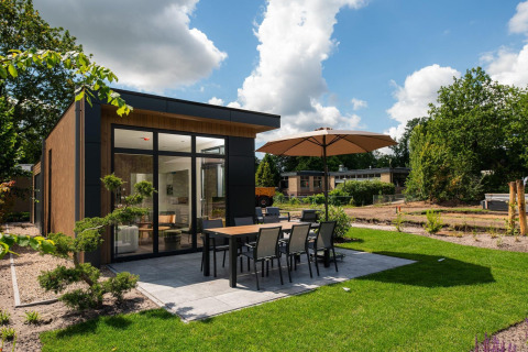 Lodge Cannenburch con patio moderno, mesa para comer, sombrilla y césped bien cuidado en un entorno natural.