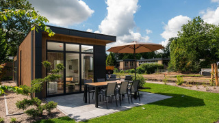 Moderne lodge Cannenburch med terrasse, spisebord, parasol og velplejet have under blå himmel.