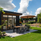 Moderne lodge Cannenburch med terrasse, spisebord, parasol og velplejet have under blå himmel.
