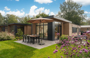 Moderne lodge med terrasse, parasol og spisebord i grøn have ved Veluwepark de Bosgraaf, Nederlandene.