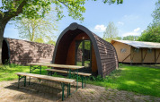Pod en Het Amsterdamse Bos, Países Bajos, rodeado de árboles y bancos de madera sobre césped verde.