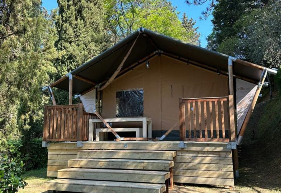 Tienda safari Villatent Wood en Camping Colleverde, Italia, rodeada de árboles y naturaleza bajo cielo azul.