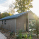 Velthorst-lodge med træbeklædning, store vinduer og en grøn have, omgivet af træer og stier, i sollys.