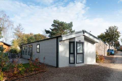 Lodge Berkel moderne avec de grandes fenêtres, dans un cadre naturel, sur sentier gravillonné sous ciel bleu.