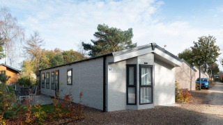 Lodge Berkel moderne avec de grandes fenêtres, dans un cadre naturel, sur sentier gravillonné sous ciel bleu.