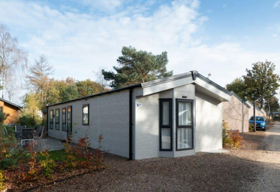 Lodge Berkel moderne avec de grandes fenêtres, dans un cadre naturel, sur sentier gravillonné sous ciel bleu.