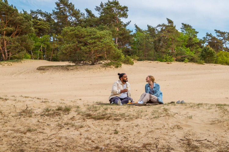 Dos personas disfrutan de un picnic sobre la arena junto a árboles en un alojamiento glamping.
